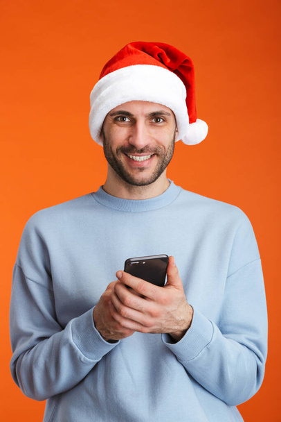 Image of a happy young man in christmas santa hat posing isolated over orange wall background holding mobile phone. - Valokuva, kuva