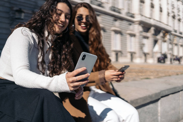 Фото на складі двох молодих жінок - козаків, які роблять самовідтворення на вулиці. Вони посміхаються. Вони носять розумний повсякденний одяг і сонячні окуляри.. - Фото, зображення