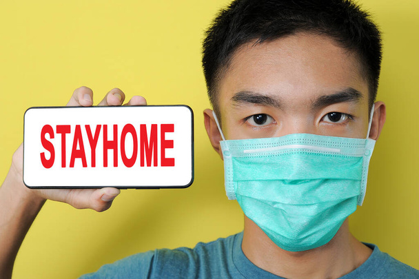 Young Asian man wearing protect mask showing STAYHOME text on phone screen beside his head, isolated on yellow background - Photo, Image