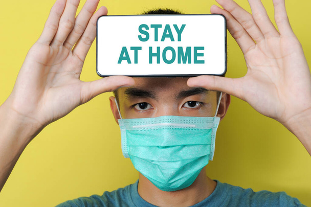 Young Asian man wearing protect mask showing STAY AT HOME text on phone screen in front of his forehead, isolated on yellow background - Zdjęcie, obraz