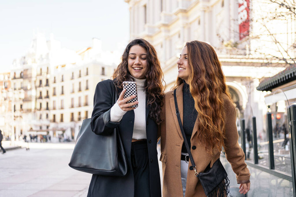 Foto en stock de dos mujeres caucásicas jóvenes usando un teléfono inteligente en la calle mientras caminan. Están sonriendo. Llevan ropa casual inteligente.
. - Foto, Imagen