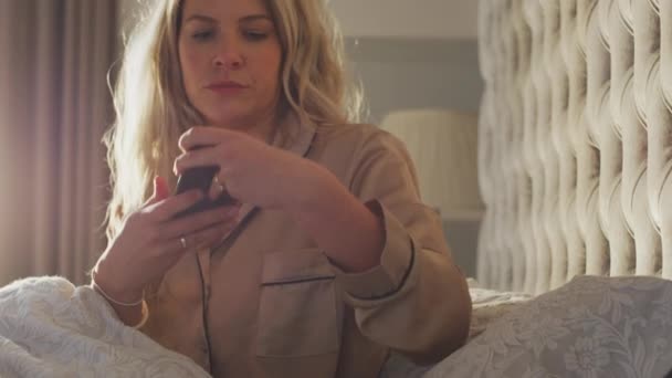 Woman getting into in bed setting sleep data app running on mobile phone on bedside table - shot in slow motion - Video, Çekim