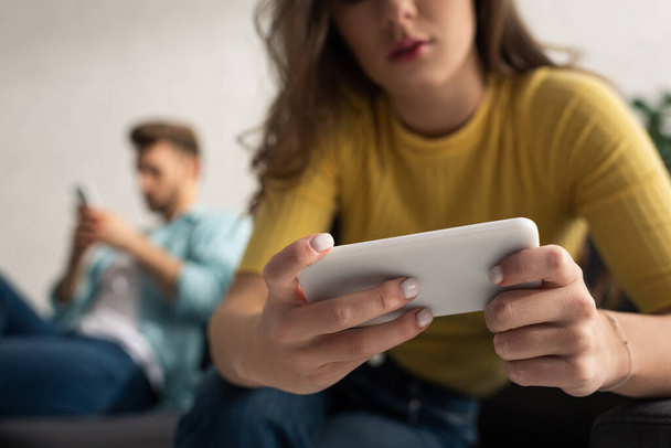 Selective focus of girl with smartphone sitting near boyfriend on couch  - Foto, afbeelding