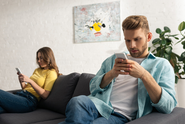Selective focus of man using smartphone near girlfriend on couch  - Valokuva, kuva