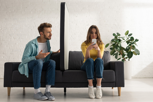 Verwirrter Mann schaut Freundin an, die in der Nähe von großem Smartphone-Modell auf Sofa chattet  - Foto, Bild