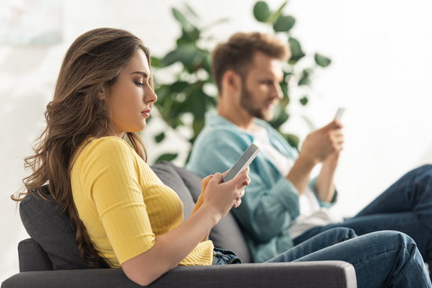 Πλευρική άποψη της νεαρής γυναίκας χρησιμοποιώντας smartphone με λευκή οθόνη κοντά φίλο στον καναπέ  - Φωτογραφία, εικόνα