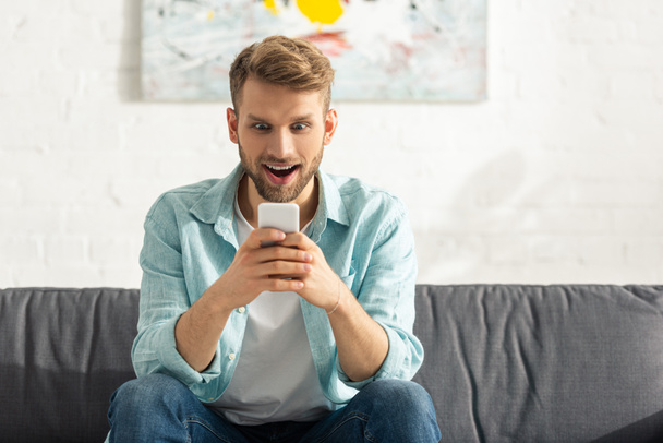 Shocked man using smartphone on couch in living room - Photo, Image