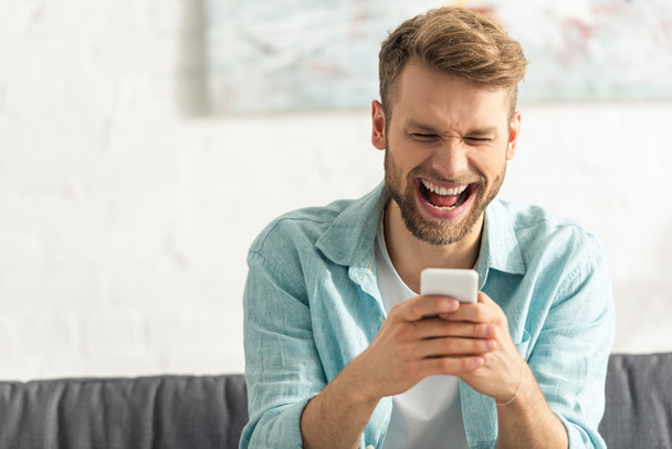 Enfoque selectivo del hombre alegre riendo mientras usa el teléfono inteligente en el sofá
  - Foto, Imagen