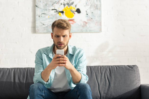 Confused man using smartphone on sofa at home  - 写真・画像