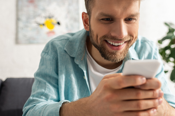Selective focus of addicted man smiling while using smartphone on couch in living room - Фото, зображення