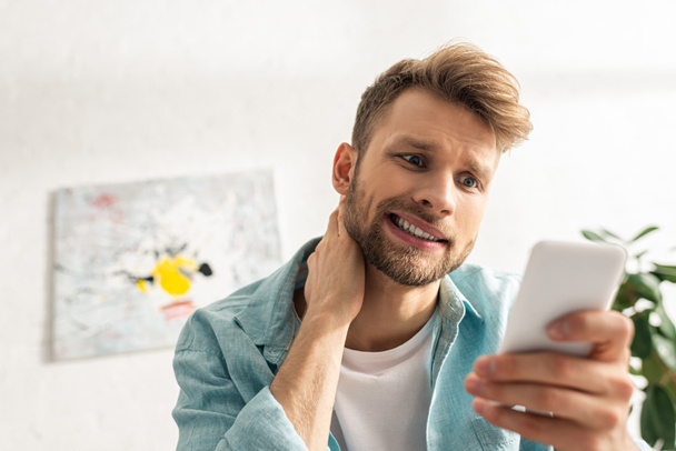 Szelektív fókusz zavarodott férfi néz smartphone a nappaliban - Fotó, kép