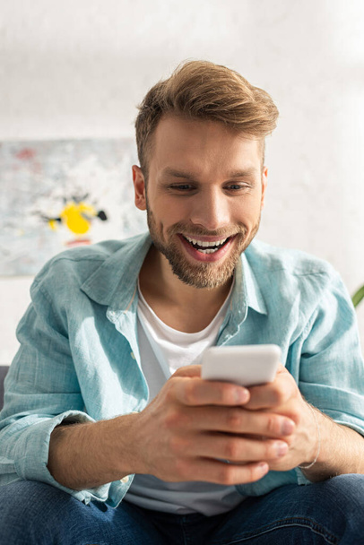 Selective focus of smiling addicted man using smartphone in living room - Fotografie, Obrázek