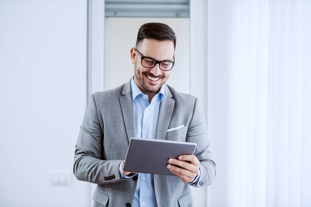 Attractive cheerful caucasian classy businessman in suit and with eyeglasses holding tablet and standing next to window. - Photo, Image