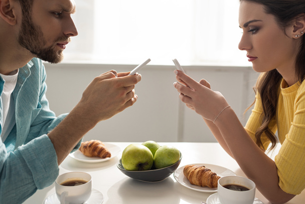 Vista lateral de la pareja utilizando teléfonos inteligentes cerca de café y cruasanes en la mesa
  - Foto, Imagen