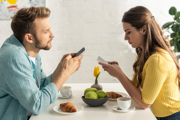 Вид збоку чоловіка в чаті біля дівчини тримає смартфон під час сніданку на кухні
  - Фото, зображення