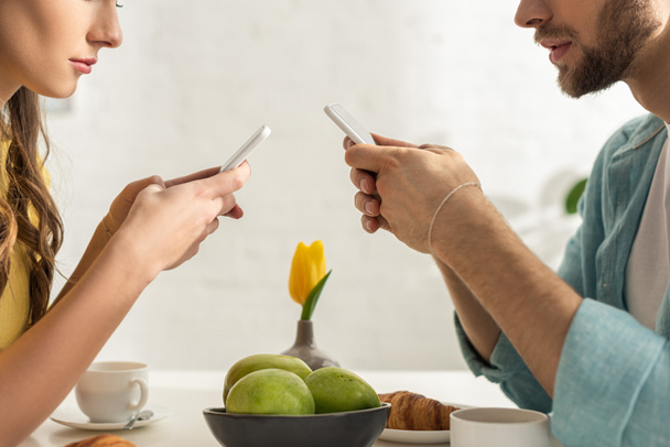 Vágott kilátás pár csevegés okostelefonok reggeli közben a konyhában  - Fotó, kép