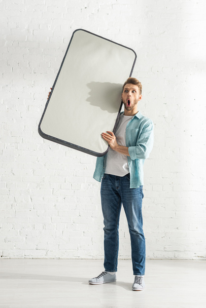 Shocked man holding model of smartphone near white brick wall  - Photo, Image
