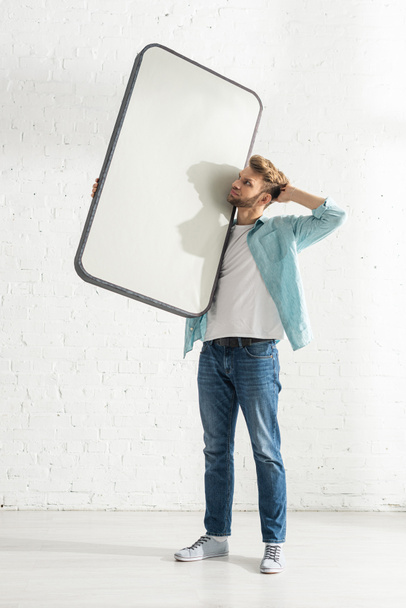 Σκεπτικός άνθρωπος με το χέρι κοντά στο κεφάλι κρατώντας το μοντέλο του smartphone στο σπίτι  - Φωτογραφία, εικόνα
