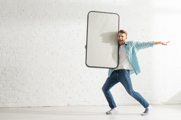 Hombre alegre riendo mientras sostiene el modelo grande de teléfono inteligente cerca de la pared de ladrillo blanco
 - Foto, Imagen