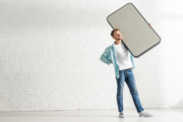 Красивий чоловік з рукою на стегні тримає велику модель смартфона біля білої цегляної стіни
 - Фото, зображення