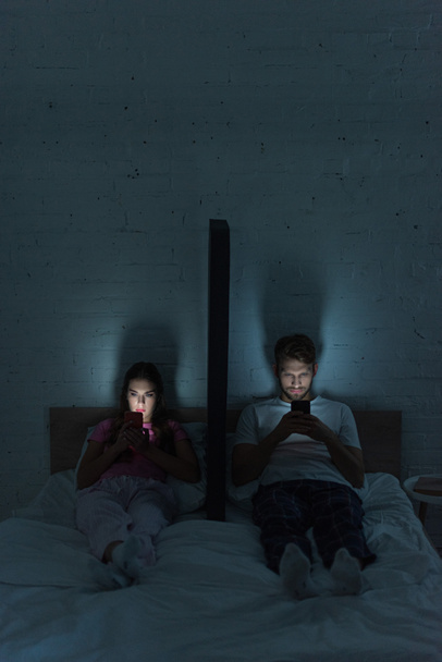 Young couple with smartphones sitting near model of smartphone on bed at night  - Foto, Imagen