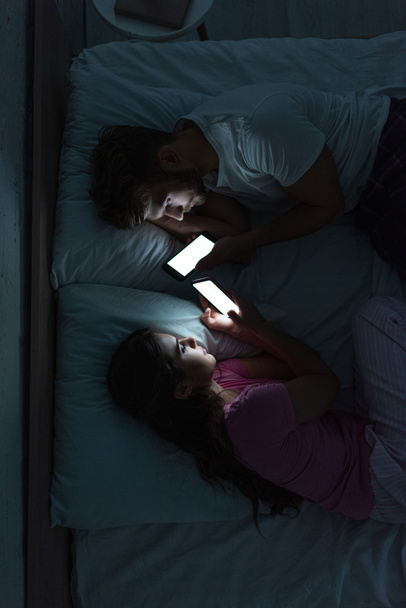 Visão superior do jovem casal dependente usando smartphones enquanto deitado na cama à noite
  - Foto, Imagem