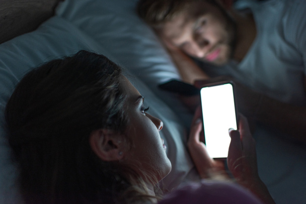 Enfoque selectivo de la chica dependiente de teléfonos inteligentes tumbado cerca de novio en la cama por la noche
  - Foto, imagen