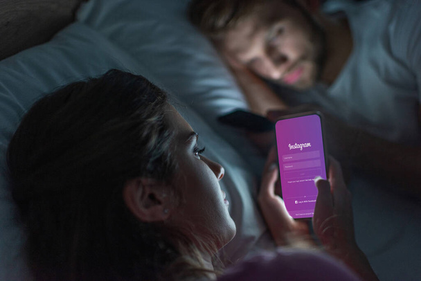 KYIV, UKRAINE - FEBRUARY 21, 2020: Selective focus of girl using smartphone with instagram app near boyfriend on bed at night  - Foto, Bild