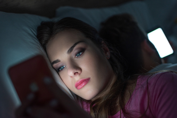 Selective focus of addicted woman using smartphone near man on bed at night  - Fotografie, Obrázek