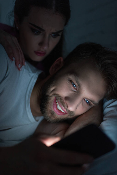 Angry woman embracing smiling boyfriend with smartphone on bed at night  - Foto, imagen