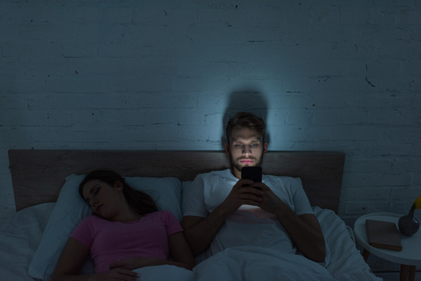 Smartphone εξαρτάται άνθρωπος κάθεται στο κρεβάτι κοντά κοιμάται φίλη  - Φωτογραφία, εικόνα