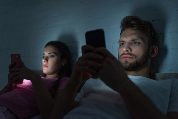 Selective focus of smartphone depended couple lying on bed at night  - Fotó, kép