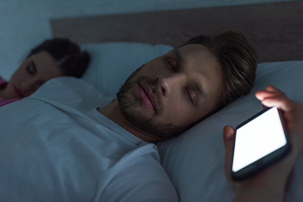 Selective focus of smartphone depended man sleeping near girlfriend on bed at night  - Fotoğraf, Görsel