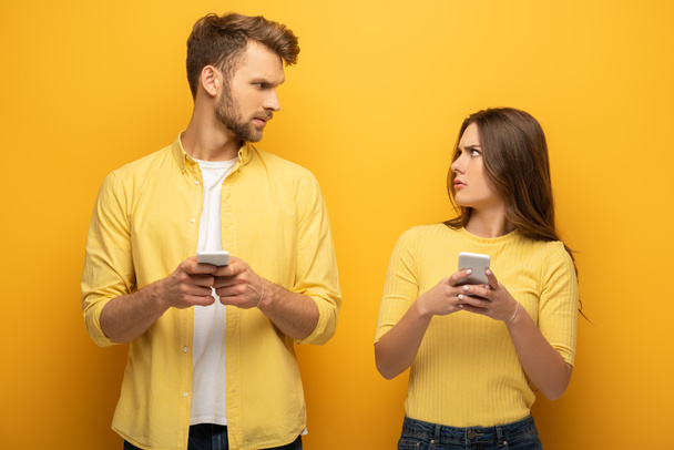 Confused couple with smartphones looking at each other on yellow background - Photo, Image