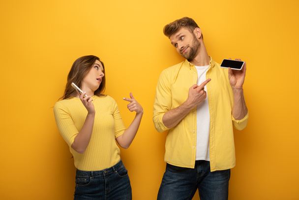 Couple pointing with fingers at smartphones and looking at each other on yellow background - Photo, Image