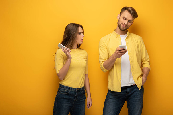 Confused girl looking at smiling boyfriend with smartphone on yellow background - Photo, Image