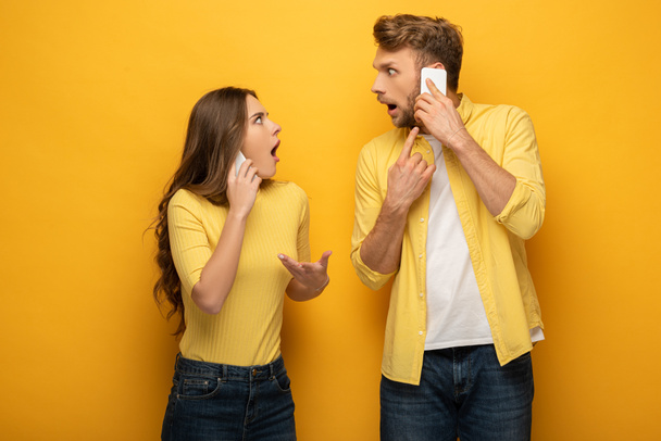 Shocked couple looking at each other while talking on smartphones on yellow background - Foto, immagini