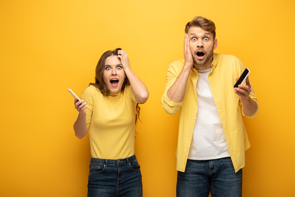 Surprised young couple holding smartphones and looking at camera on yellow background - Fotografie, Obrázek