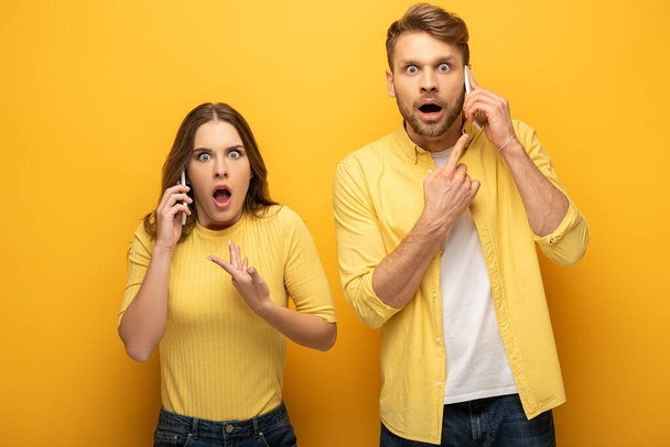 Überraschtes Paar zeigt mit dem Finger, während es auf Smartphones auf gelbem Hintergrund spricht - Foto, Bild