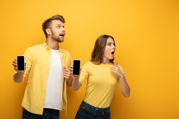 Šokovaný mladý pár dívá pryč, zatímco ukazuje smartphony s prázdnými obrazovkami na žlutém pozadí - Fotografie, Obrázek