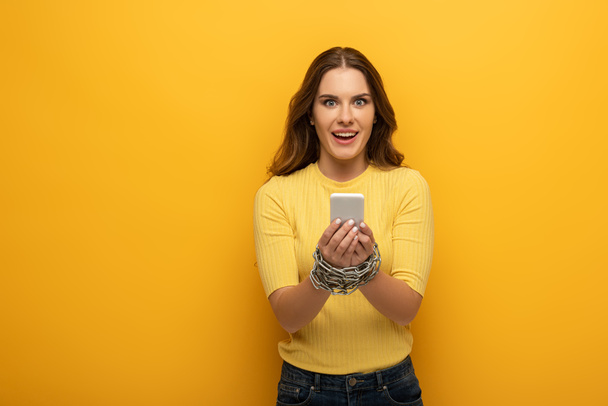 Mulher sorridente bonita com as mãos amarradas com corrente de metal segurando smartphone no fundo amarelo
 - Foto, Imagem