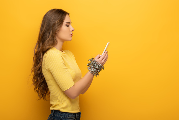 Zijaanzicht van aantrekkelijke vrouw met smartphone in gebonden handen met metalen ketting op gele achtergrond - Foto, afbeelding
