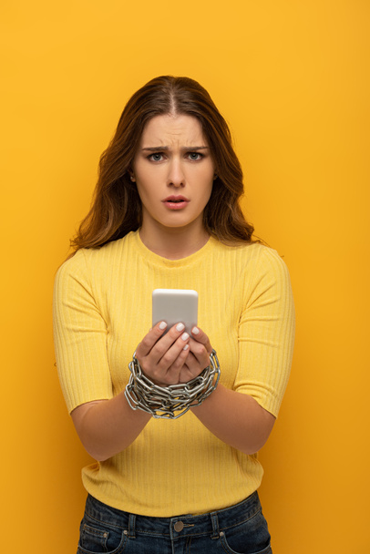Confused girl looking at camera while holding smartphone in tied hands with metal chain on yellow background - Foto, Imagem