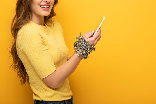 Cropped view of smiling woman holding smartphone in tied hands with chain on yellow background - 写真・画像