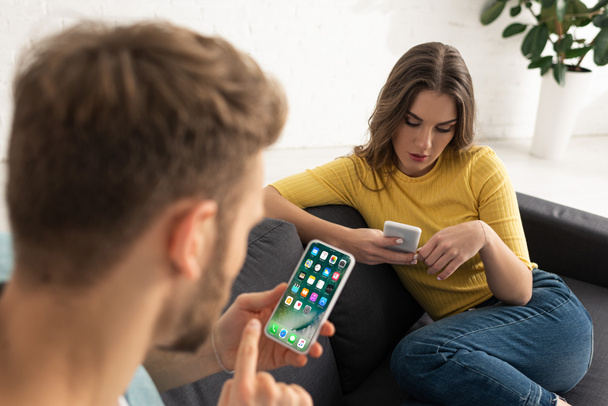 KYIV, UKRAINE - FEBRUARY 21, 2020: Selective focus of man using smartphone with iphone screen near girlfriend chatting on sofa - Фото, изображение