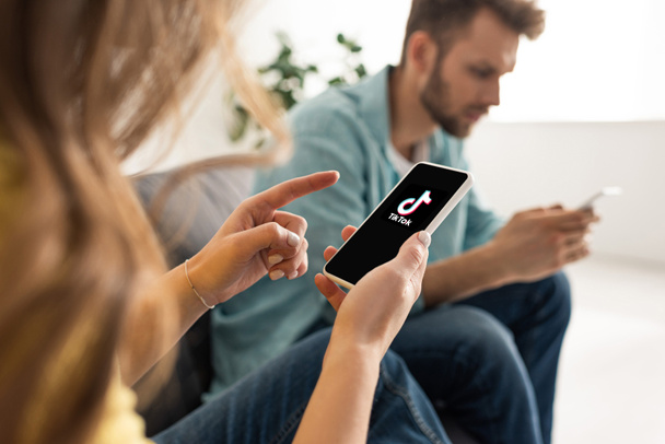 KYIV, UKRAINE - FEBRUARY 21, 2020: Selective focus of woman pointing with finger at smartphone with TikTok app near boyfriend chatting on couch  - Φωτογραφία, εικόνα