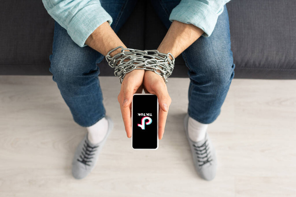 KYIV, UKRAINE - FEBRUARY 21, 2020: Top view of man with metal chain around hands holding smartphone with TikTok app on couch  - Фото, изображение