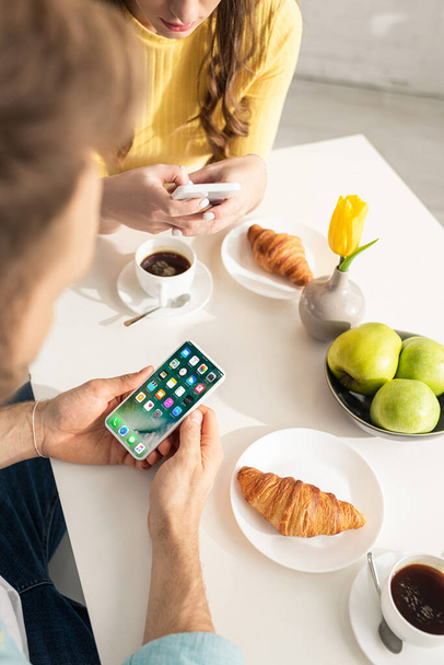 KYIV, UKRAINE - FEBRUARY 21, 2020: Selective focus of man holding smartphone with iphone screen near girlfriend and breakfast in kitchen  - Photo, Image