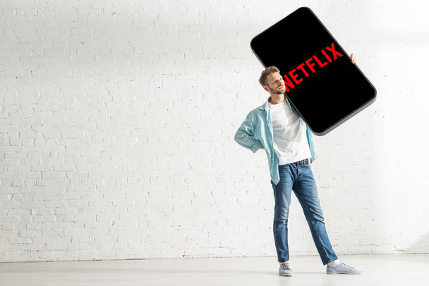 KYIV, UKRAINE - FEBRUARY 21, 2020: Handsome man with hand on hip holding big model of smartphone with netflix app at home  - Фото, зображення