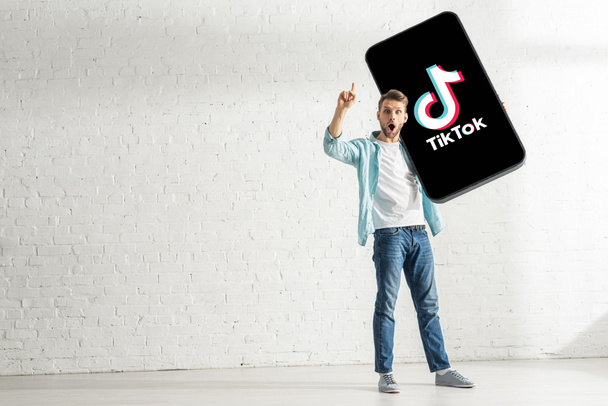 KYIV, UKRAINE - FEBRUARY 21, 2020: Surprised man showing solution gesture while holding model of smartphone with TikTok app at home  - Fotoğraf, Görsel